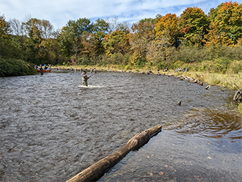 River curving up and to the left. A person is fishing in the middle and several people in a canoe are rounding the bend at the back-left